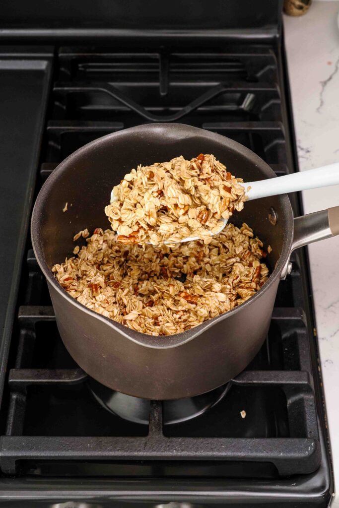 A spatula holds a spoonful of mixed granola in a pot over the stove.
