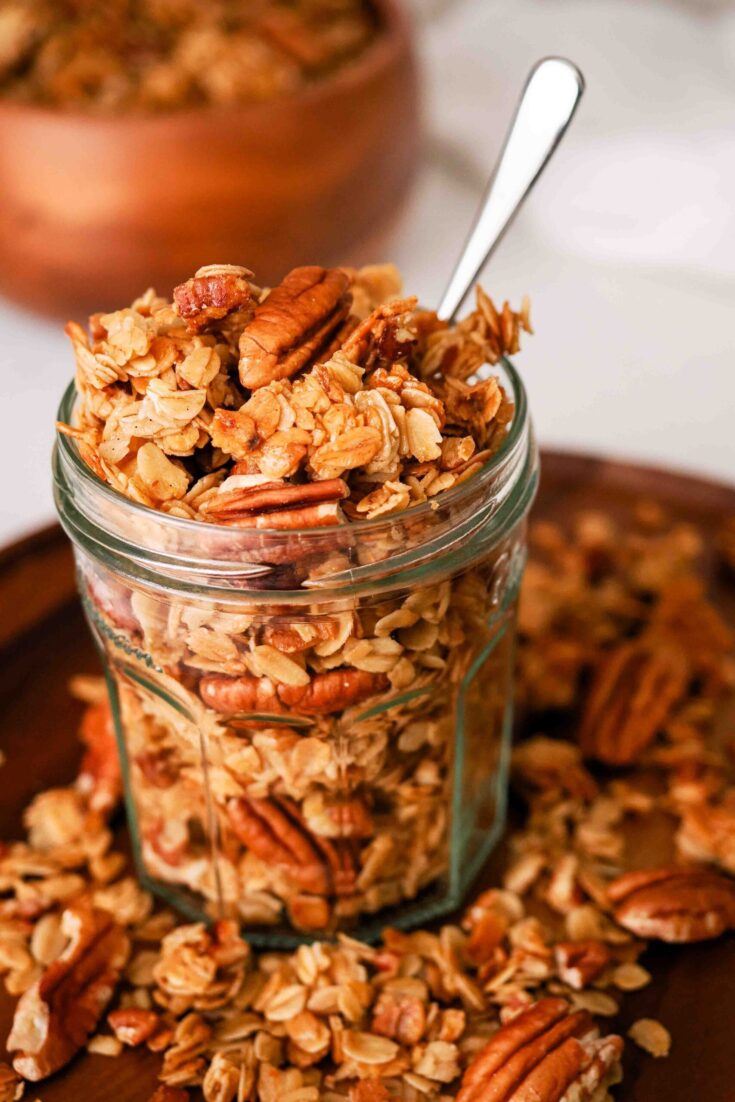 A glass jar with a small spoon sticking out is filled with honey pecan granola.