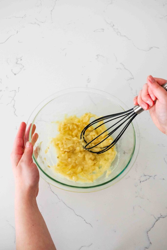 A hand mashes bananas with a large silicone whisk in a glass bowl.