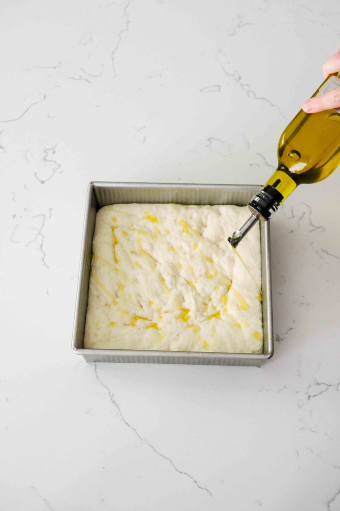 A bottle of olive oil drizzles oil over a small batch of focaccia dough.