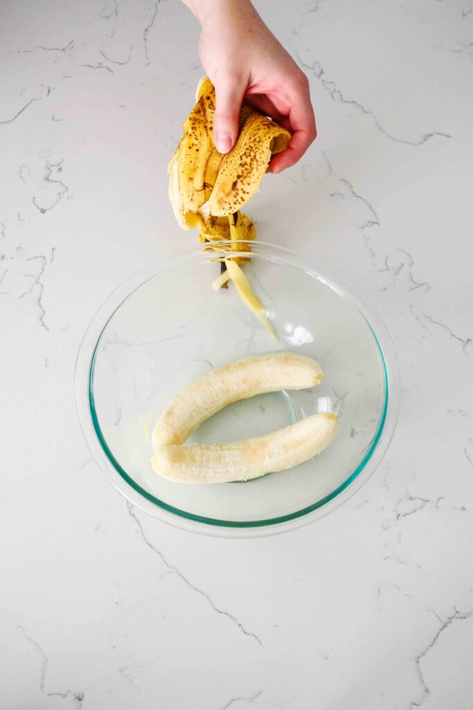 A hand holds two banana peels over a glass mixing bowl with two bananas in it.