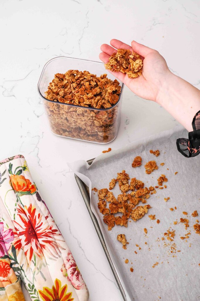 A hand pours small granola clusters into a clear reusable container.