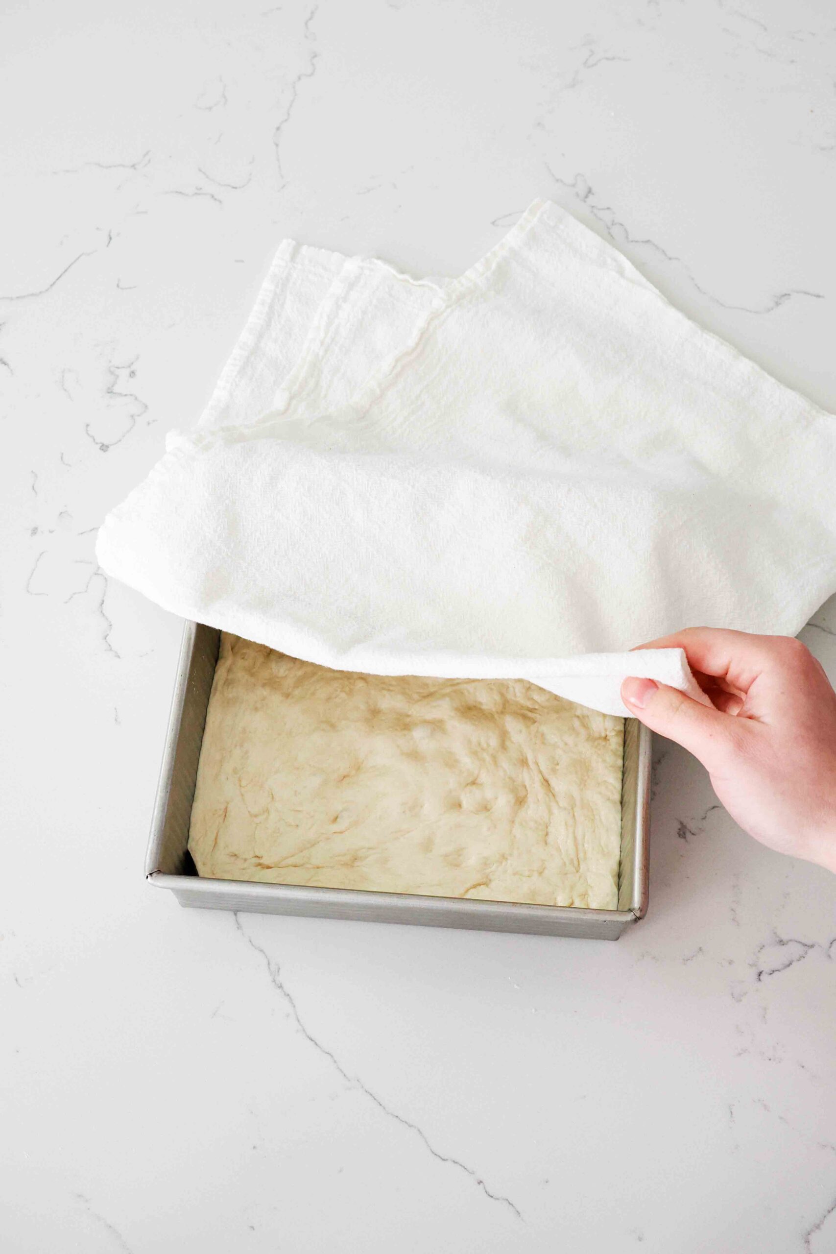 A hand pulls a kitchen towel over a square pan with focaccia dough in it.