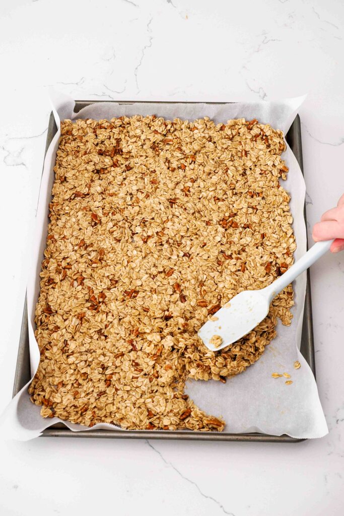 A spatula spreads granola across a parchment-lined baking sheet.
