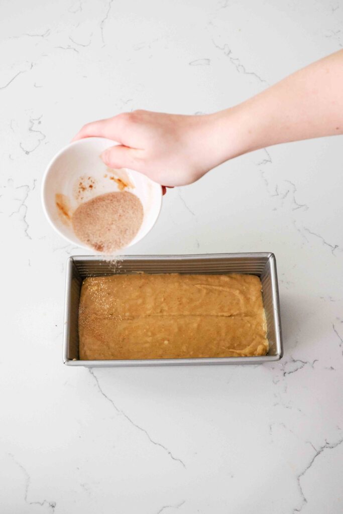 A hand sprinkles cinnamon sugar over cinnamon banana bread batter in a loaf pan.