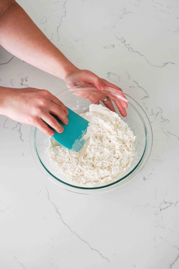 A hand uses a flexible bench scraper to stir together flour, water, yeast, and salt.