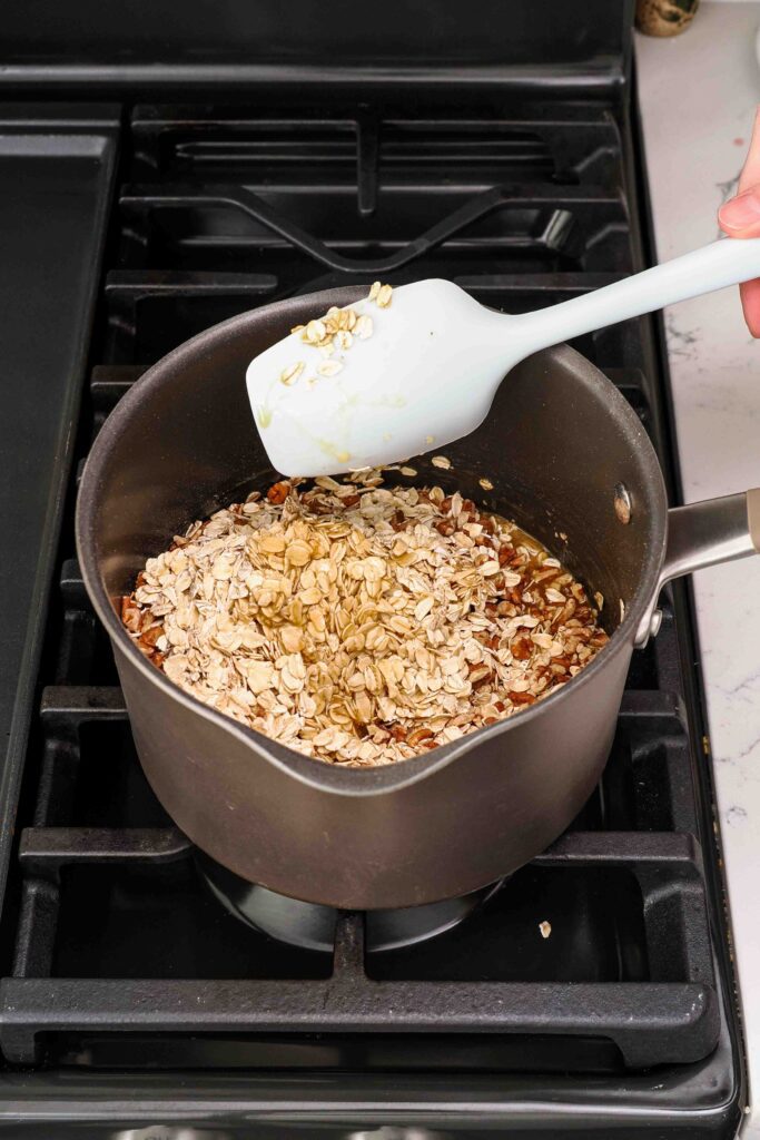 A spatula stirs rolled oats into a melted honey syrup.