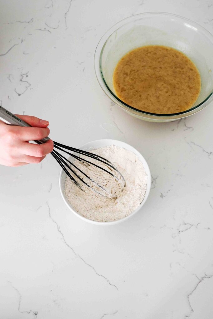 A hand whisks flour, cinnamon, salt, and baking soda together in a medium bowl.