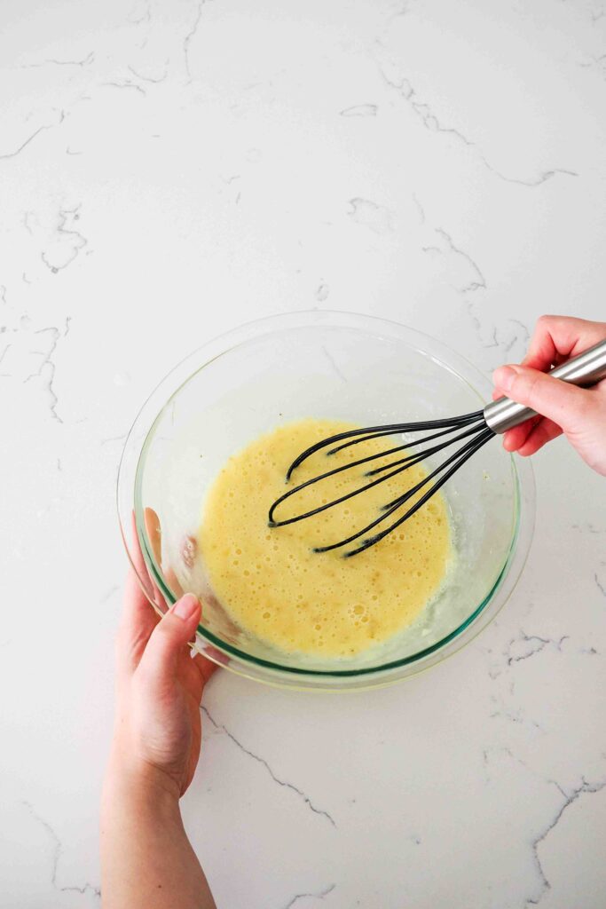 A hand whisks eggs and mashed bananas together in a glass mixing bowl.