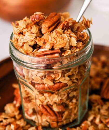 A glass jar with a small spoon sticking out is filled with honey pecan granola.