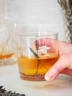 A hand picks up a rocks glass filled with a lavender old fashioned.