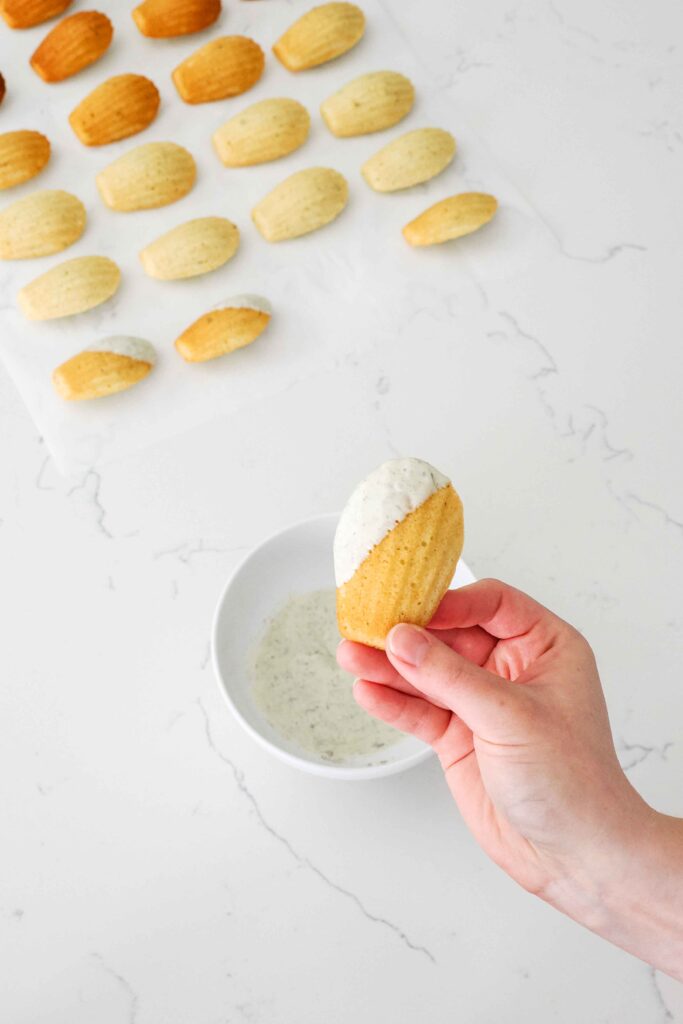 A hand holds up a partially dipped lavender madeleine with a lavender white chocolate coating.
