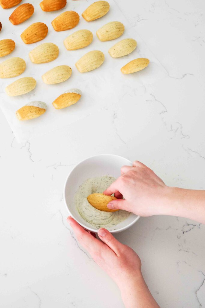 A hand dips a lavender madeleine at an angle into lavender white chocolate in a small bowl.