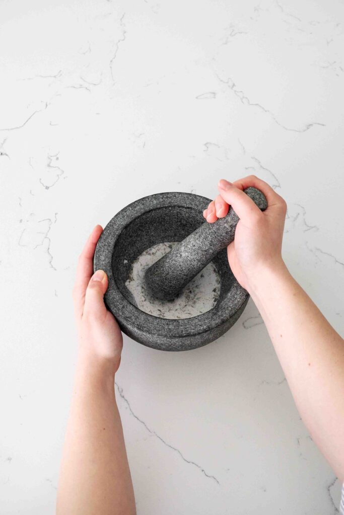 Two hands grind lavender and granulated sugar in a mortar and pestle.
