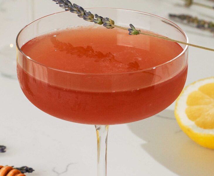 A dark pink cocktail in a coupe glass with a lavender sprig garnish.
