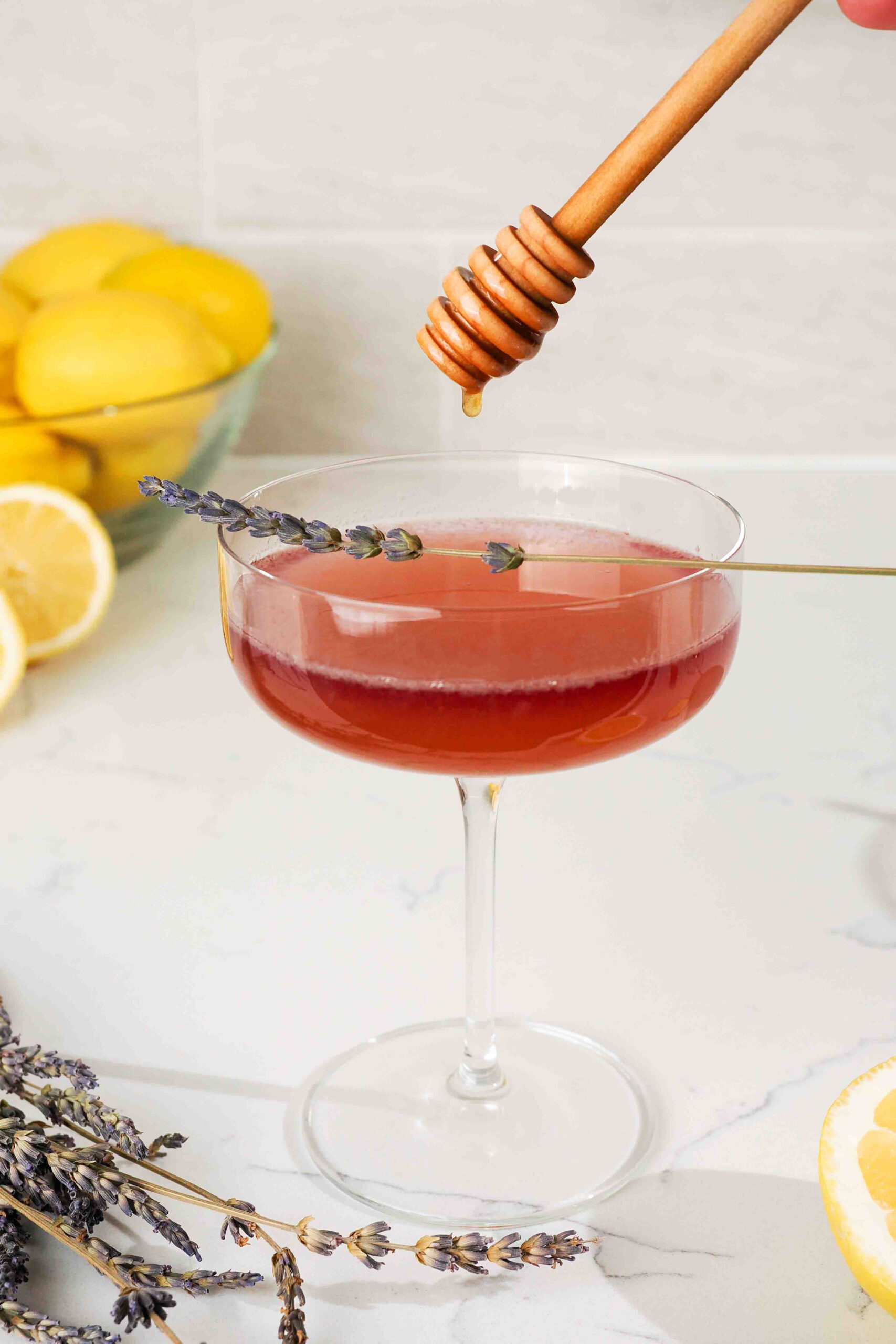 A honey dipper over a dark pink lavender bee's knees cocktail on a quartz counter.