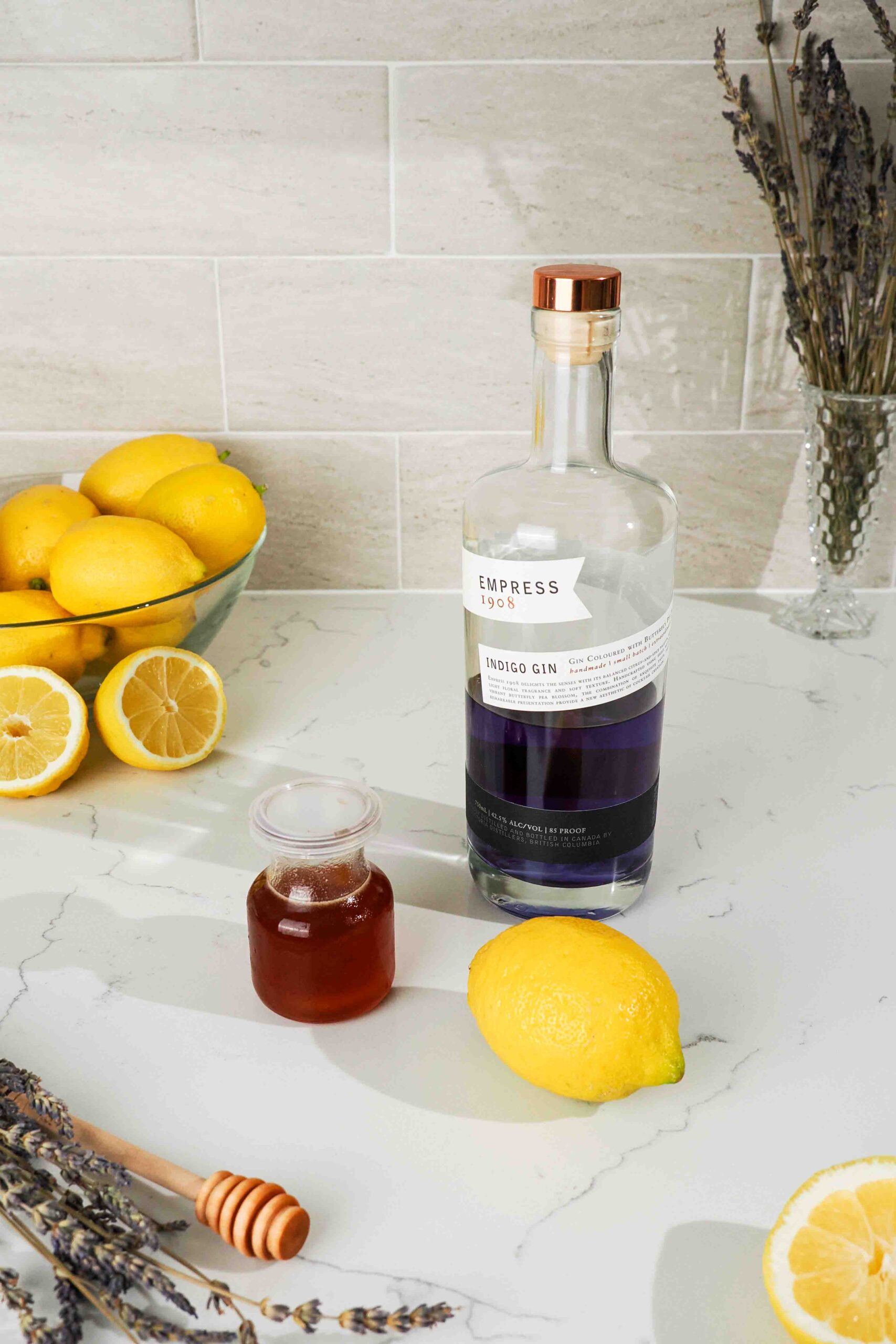 A bottle of botanical gin, honey lavender syrup, and a lemon on a quartz counter.