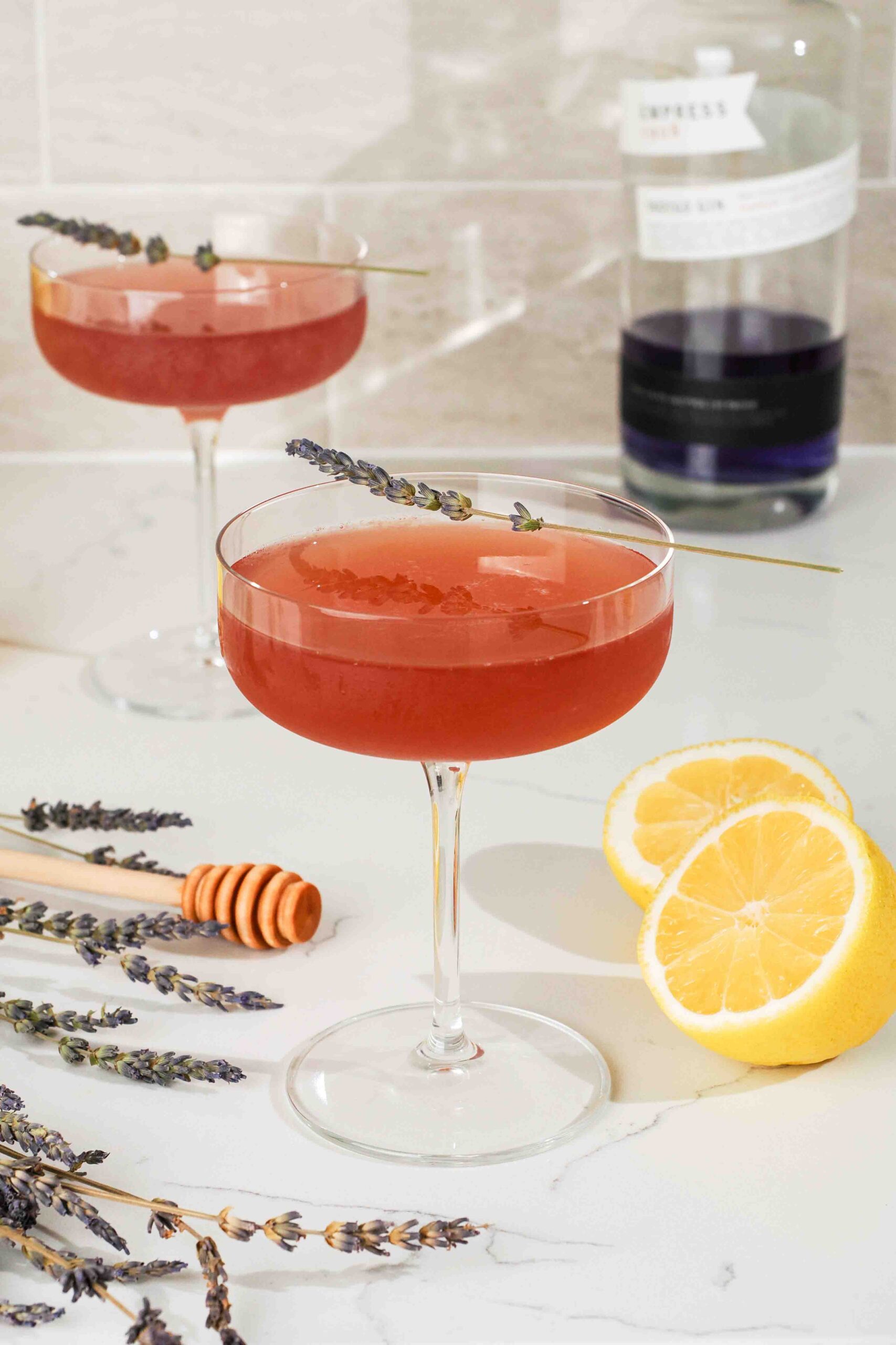 Two dark pink cocktails by lemons, lavender, and a honey dipper.