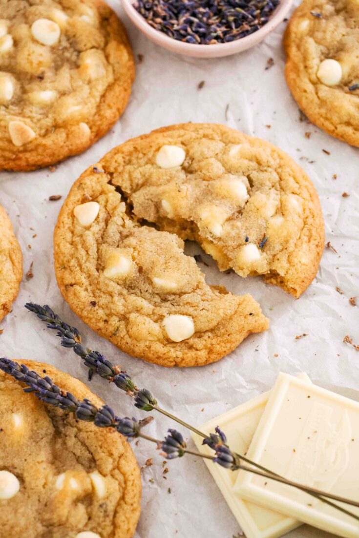 A lavender chocolate chip cookie broken in half on crumpled parchment paper with two sprigs of lavender and white chocolate pieces nearby.