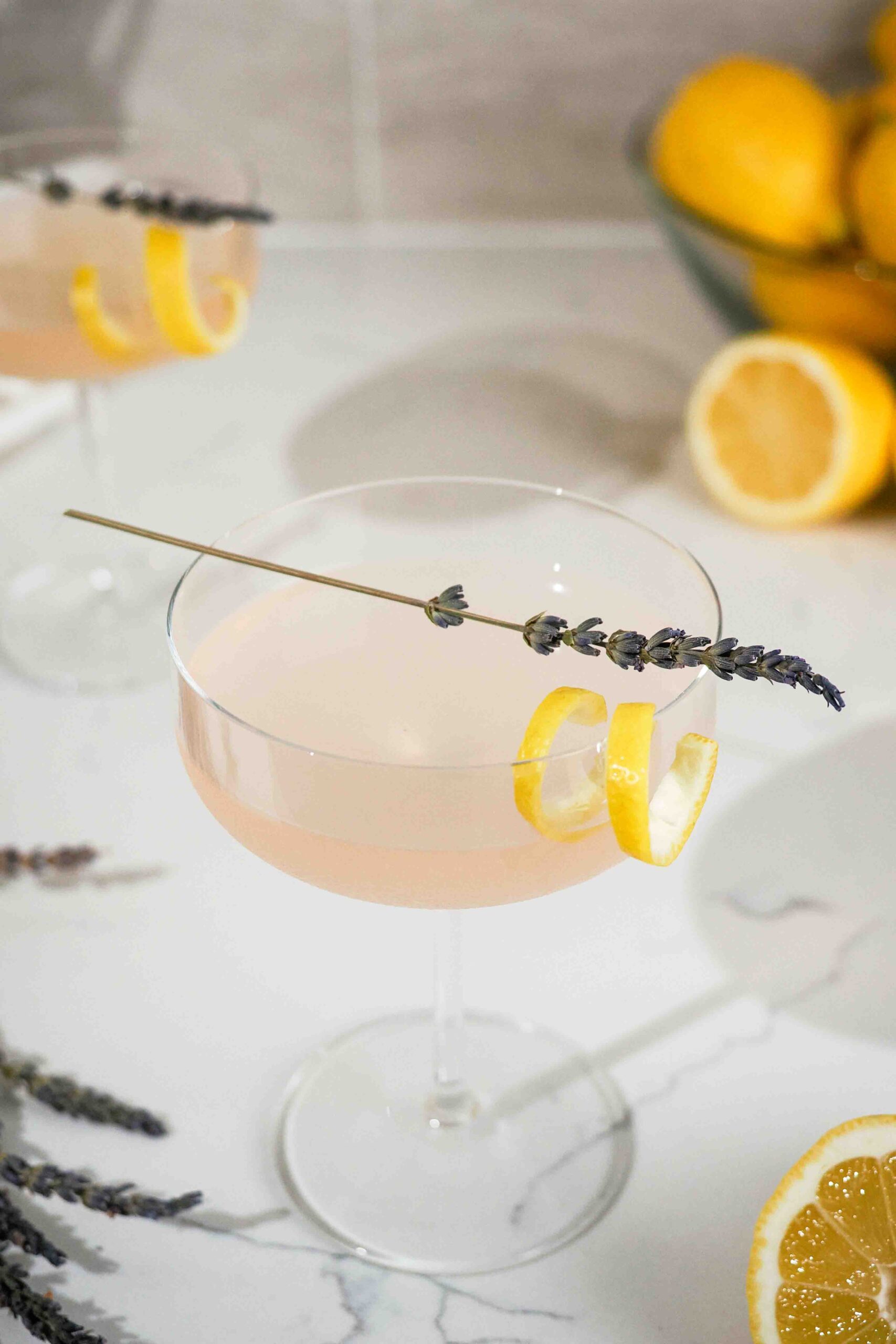 A lavender lemon drop martini in a coupe glass on a counter with lemons.