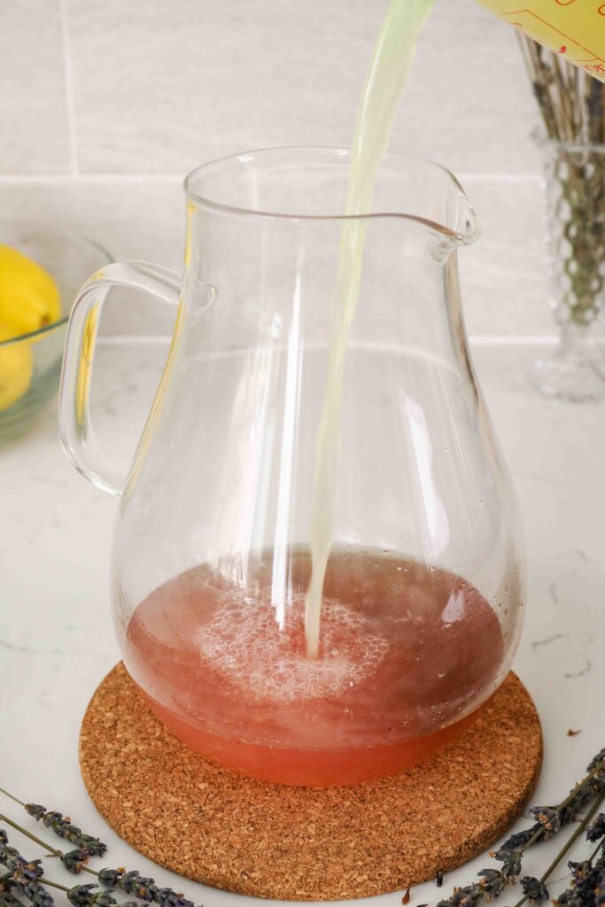 Lemon juice is poured into a pitcher with lemon lavender simple syrup. The simple syrup changes color from a dark purple to a bright pink.