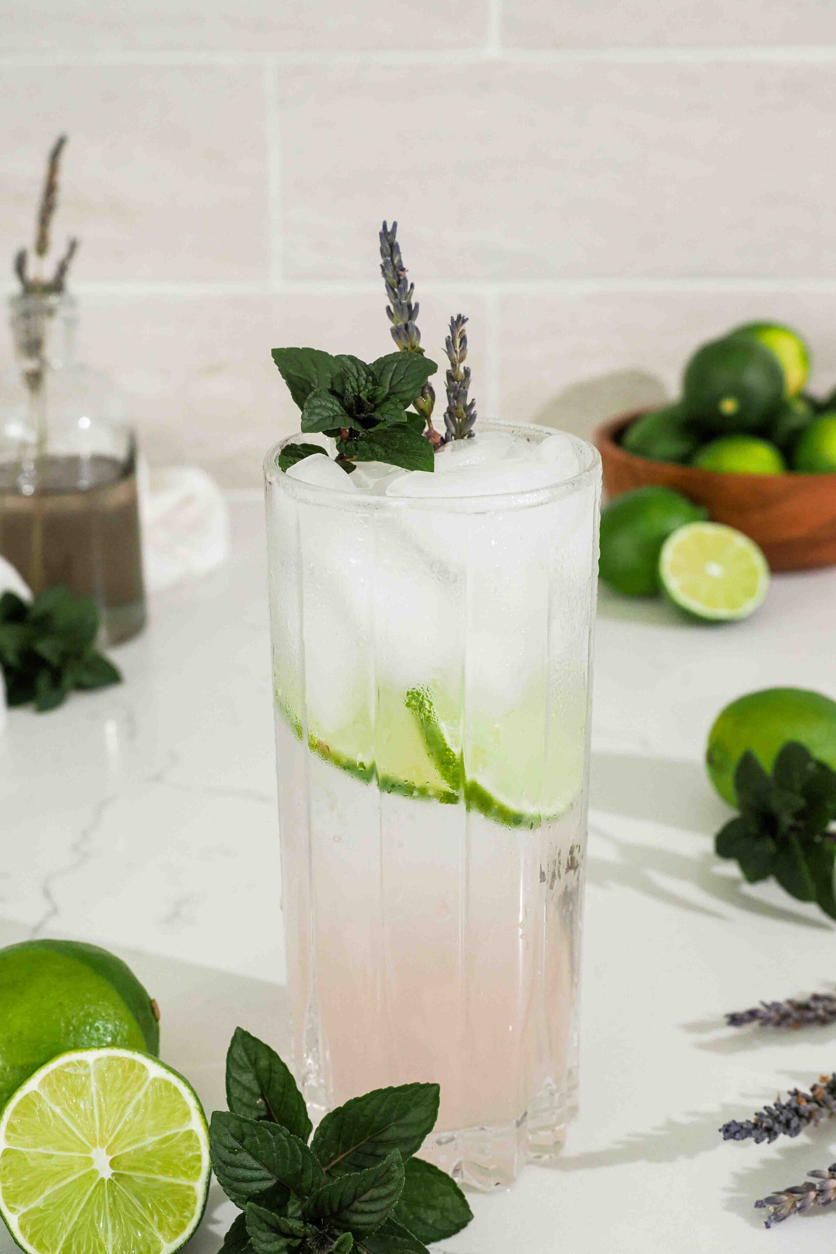 A closeup of a pink lavender mojito cocktail on a counter with limes and mint leaves.