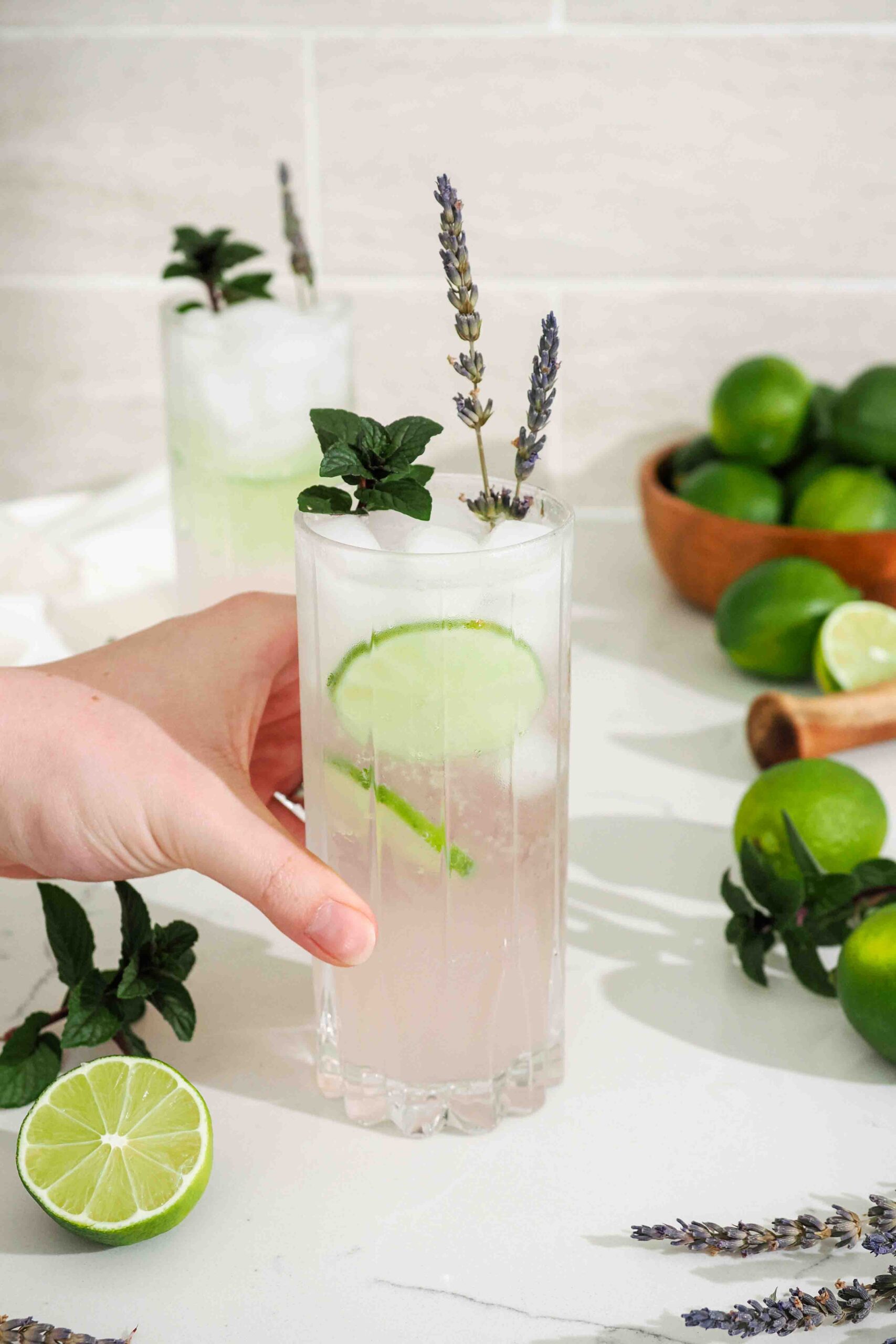 A hand holds a pink lavender mojito with mint, lime, and lavender garnishes.