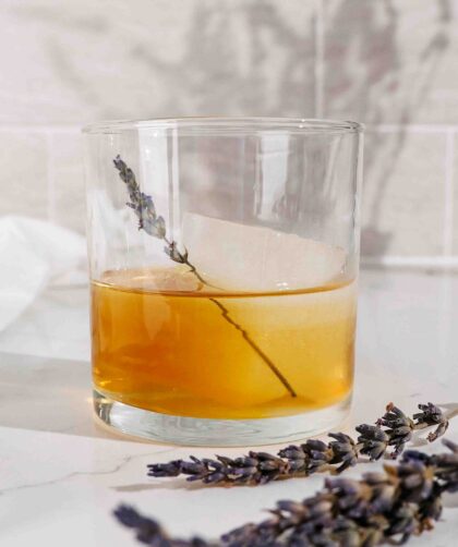 A rocks glass filled with a large ice cube and lavender old fashioned cocktail.