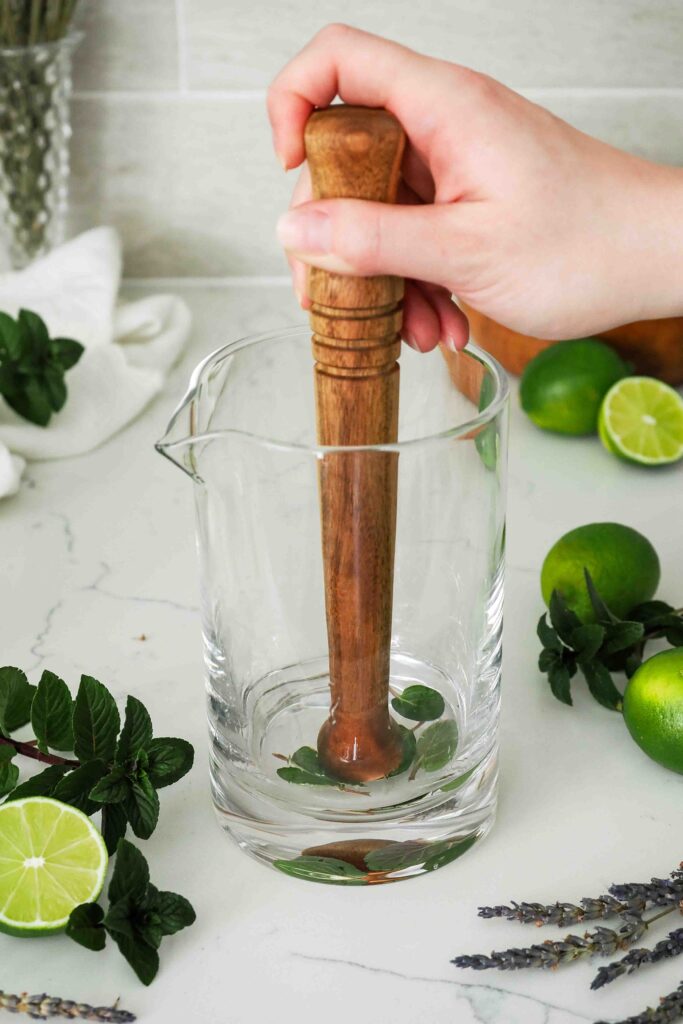 A hand uses a wooden muddler to muddle mint in white rum in a mixing glass.