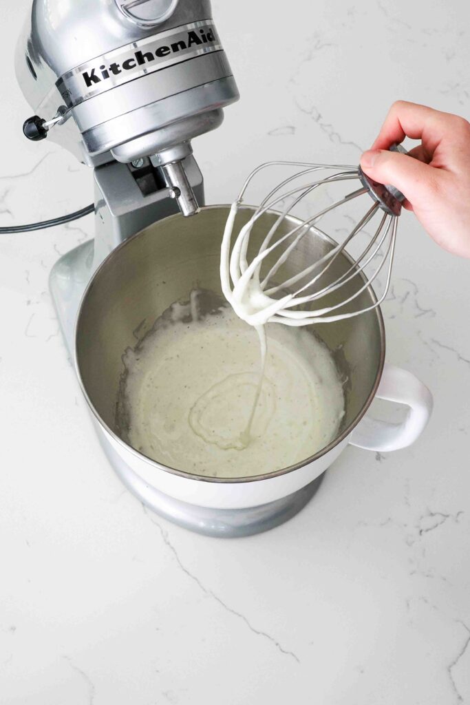 A hand holds up a whisk with pale yellow, almost white batter that leaves a ribbon on the batter in the bowl below it.