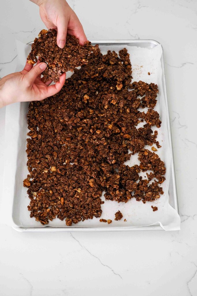 Two hands break a large slab of dark chocolate granola into smaller pieces.