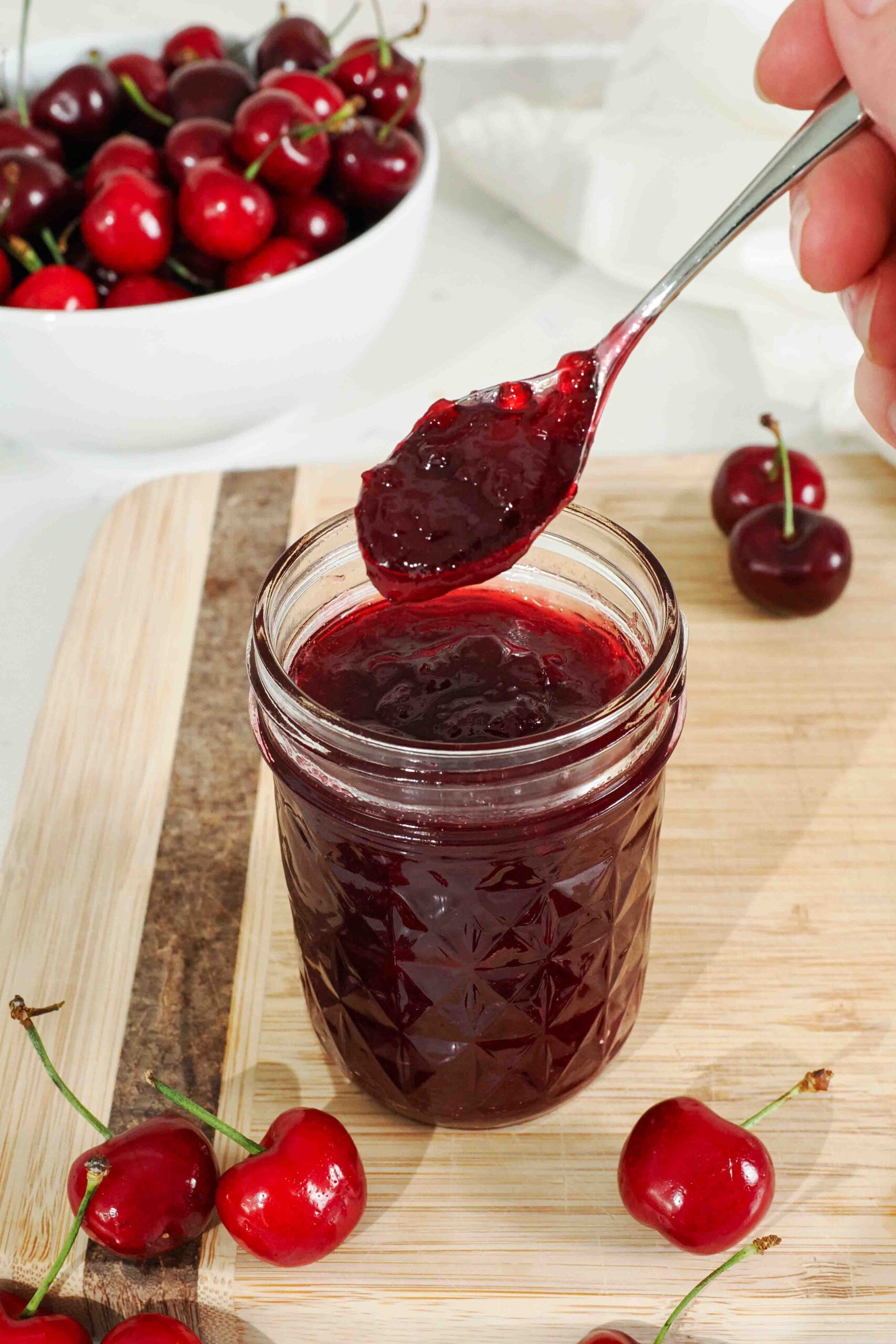 A hand holds up a spoonful of cherry jam from a glass jar.