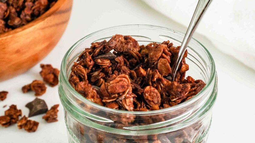A spoon sticks out of a glass jar filled with chocolate granola.