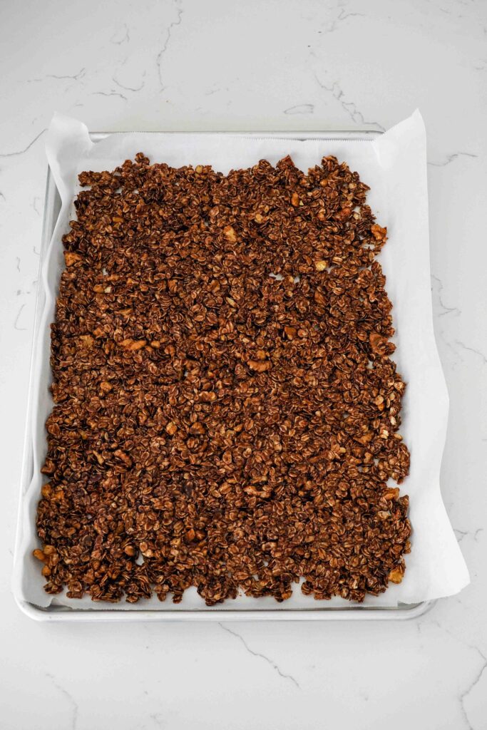 Dark chocolate granola is spread thinly in a parchment-lined half-sheet pan.