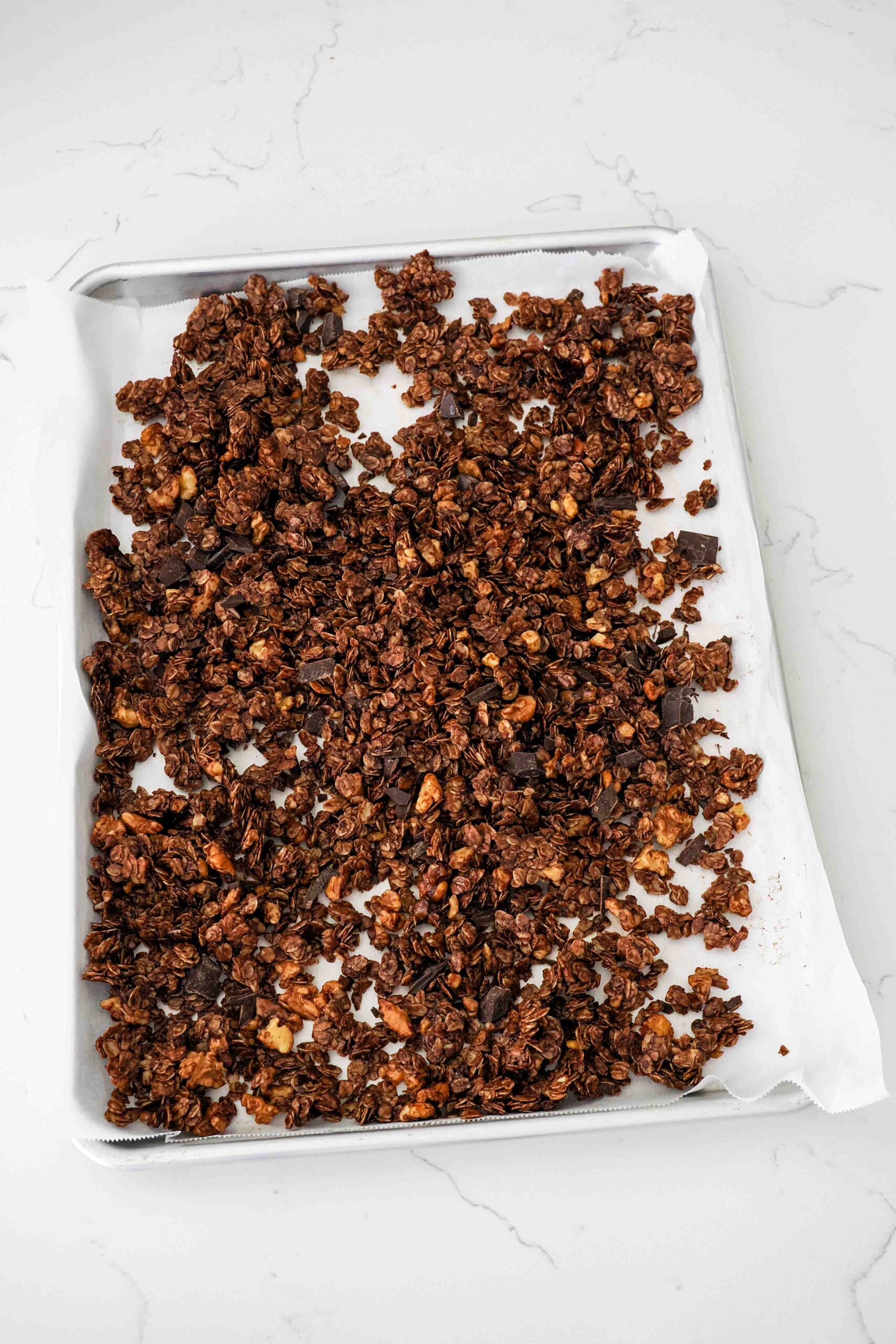 Dark chocolate granola mixed with dark chocolate chunks on a lined baking sheet.