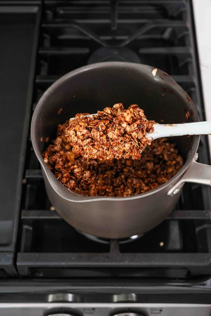 A spatula holds up dark chocolate granola over a pot on the stove.