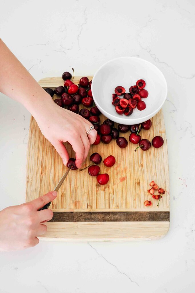 Two hands slice cherries in half on a cutting board with a paring knife.