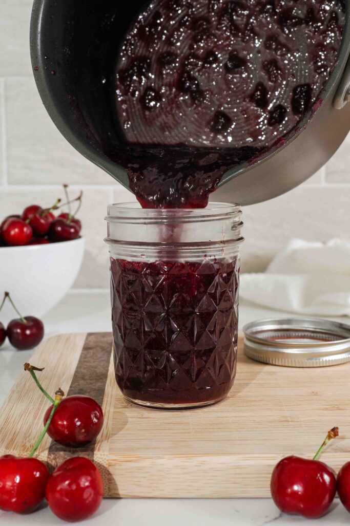 Cherry jam is poured into a half-pint glass jar.