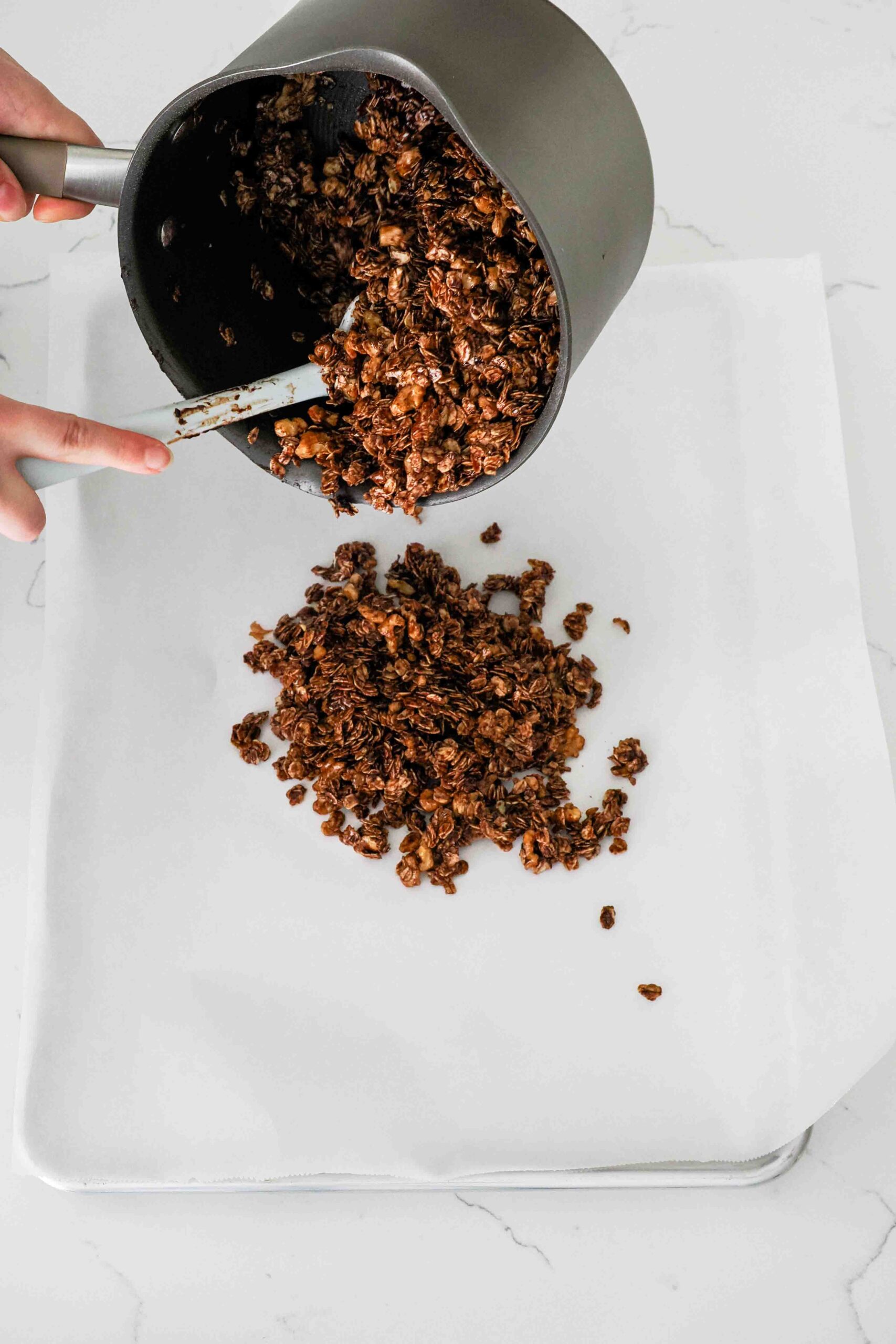 A spatula guides granola out of a pot and onto a lined baking sheet.