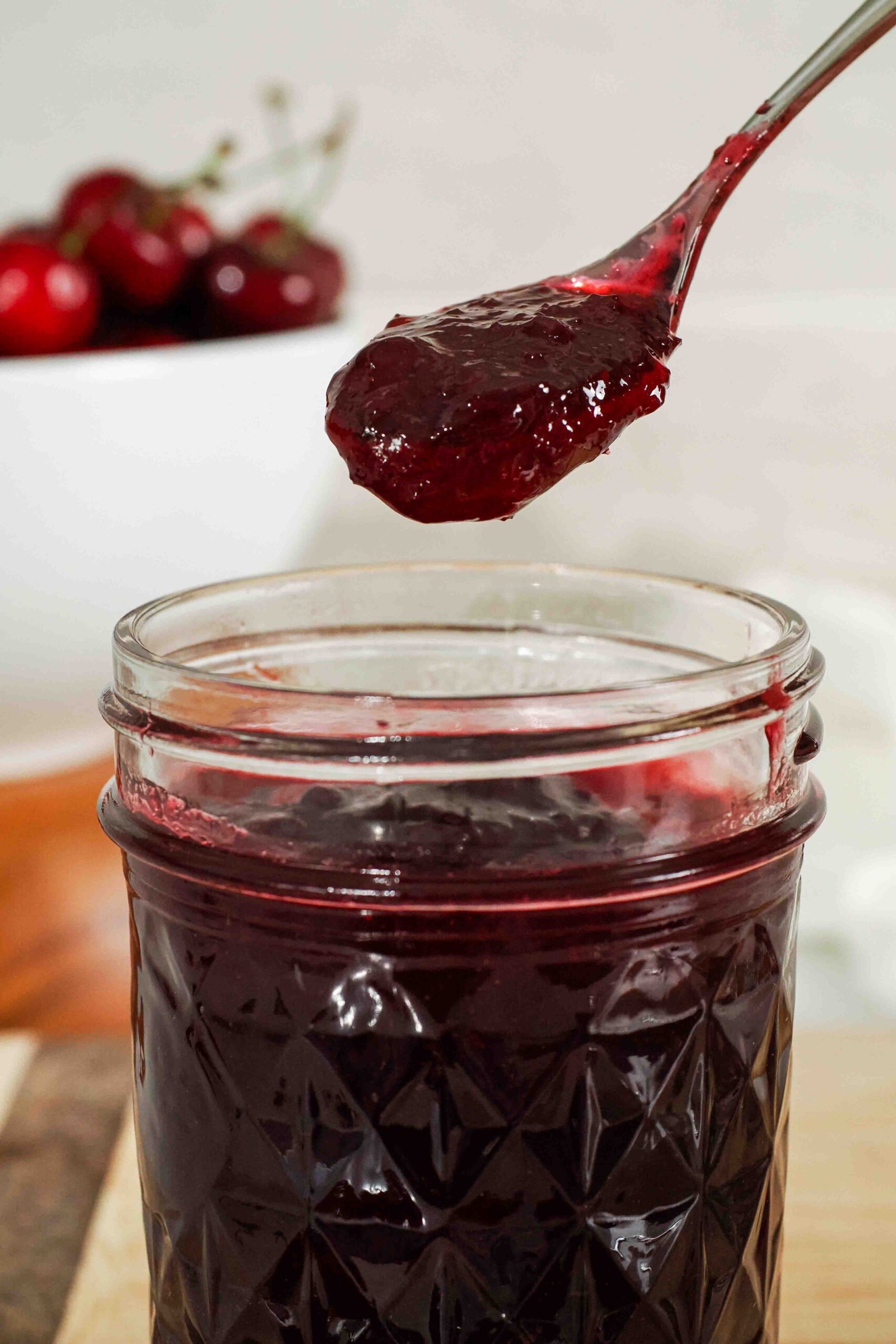 A spoon holds up chunky cherry jam over a jar.
