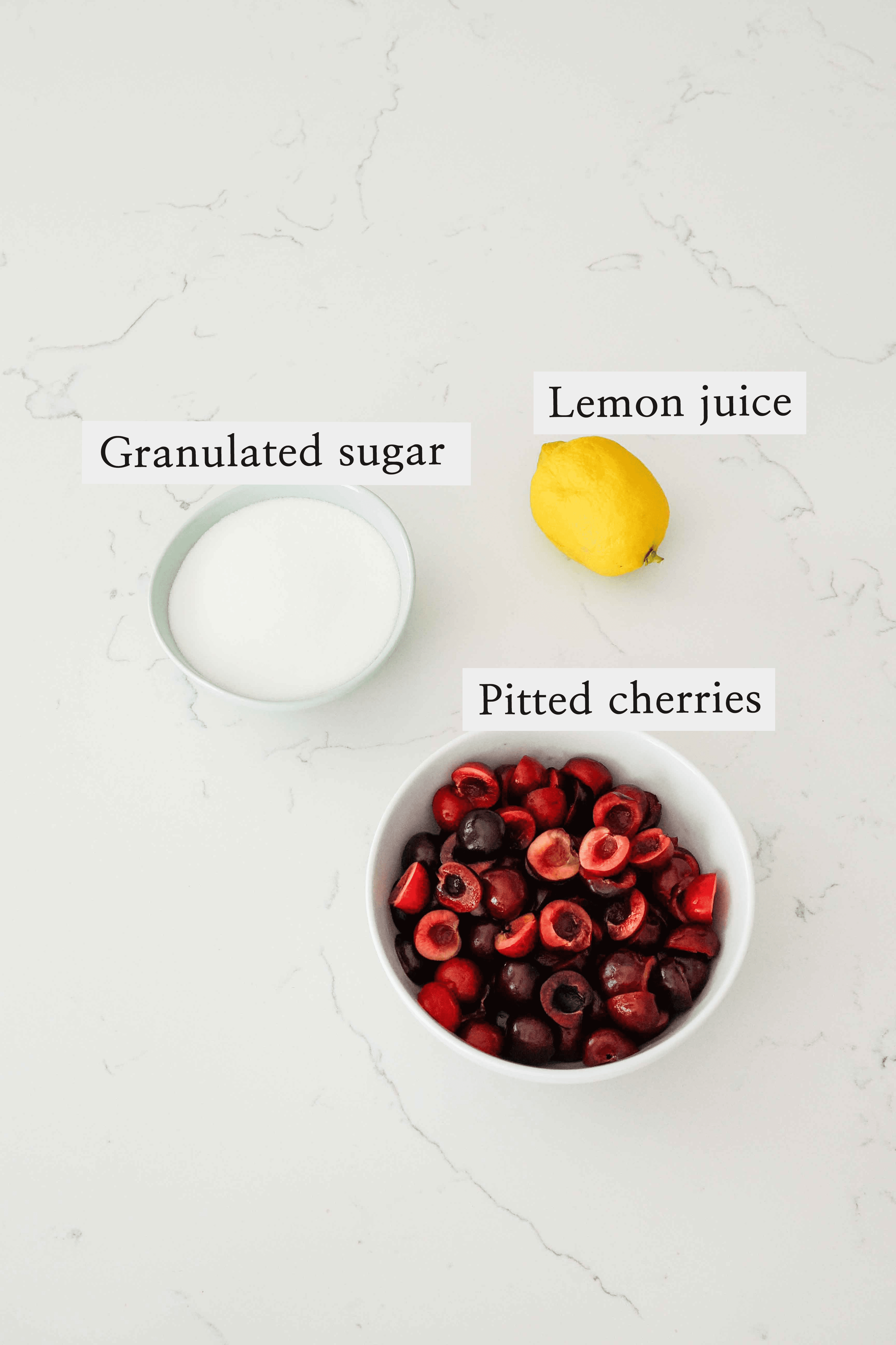 A bowl of halved cherries, a lemon, and a bowl of sugar on a white quartz counter.
