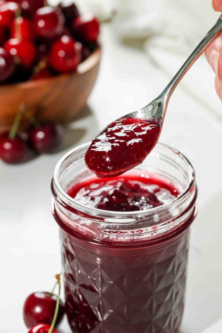A small spoon scoops cherry jam out of a half-pint jar.