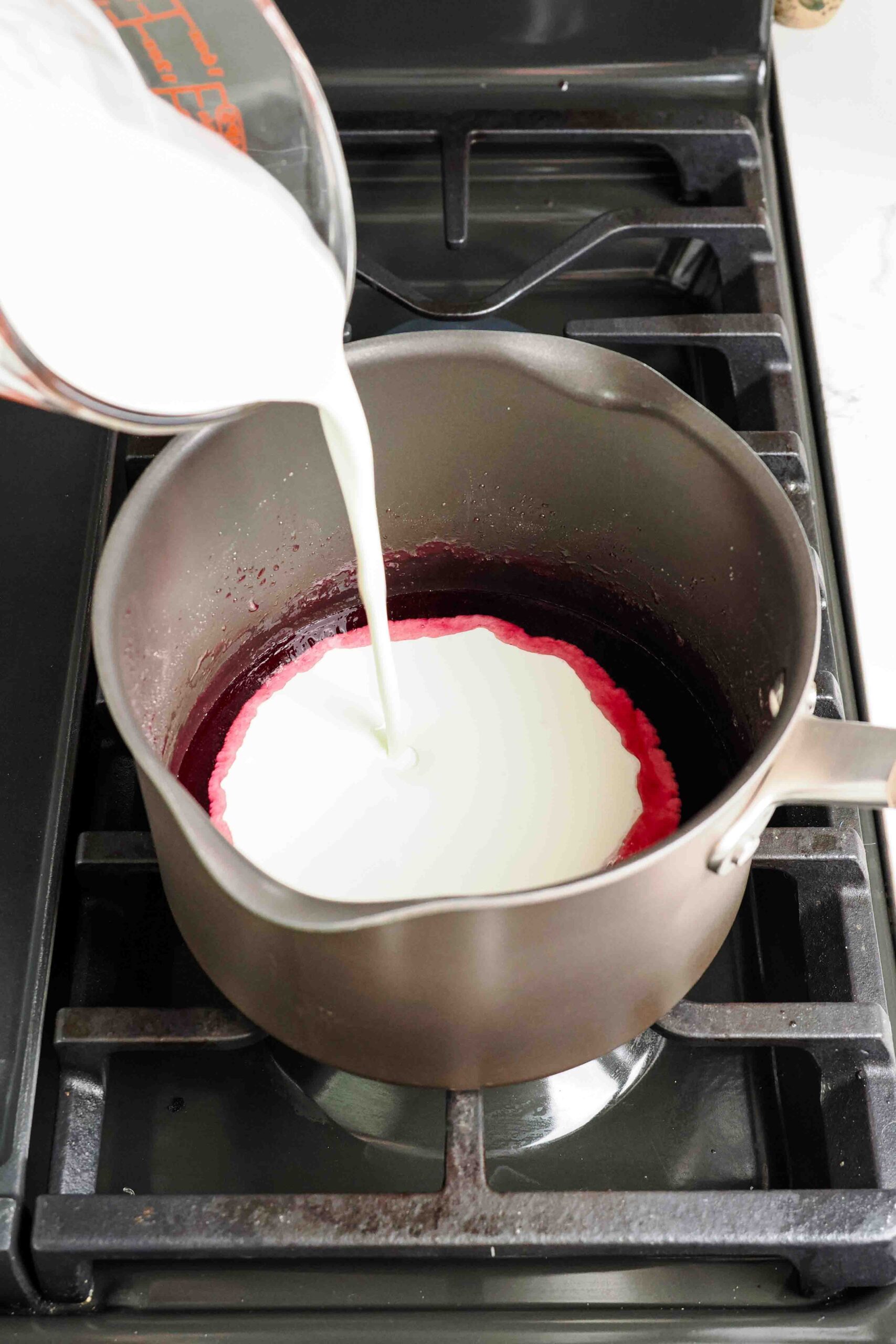 A measuring cup pours heavy cream into cooled cherry syrup on the stove.
