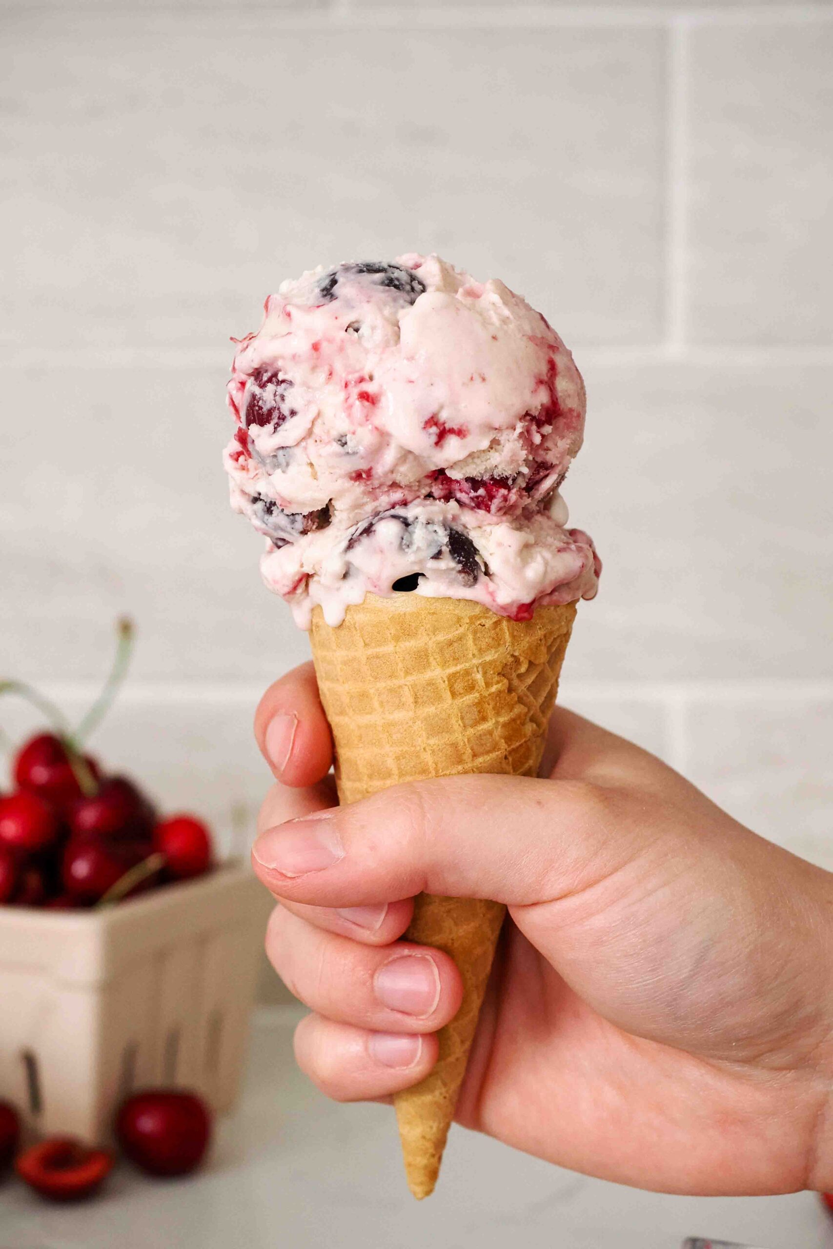 A hand holds a double scooped cone of black cherry ice cream.
