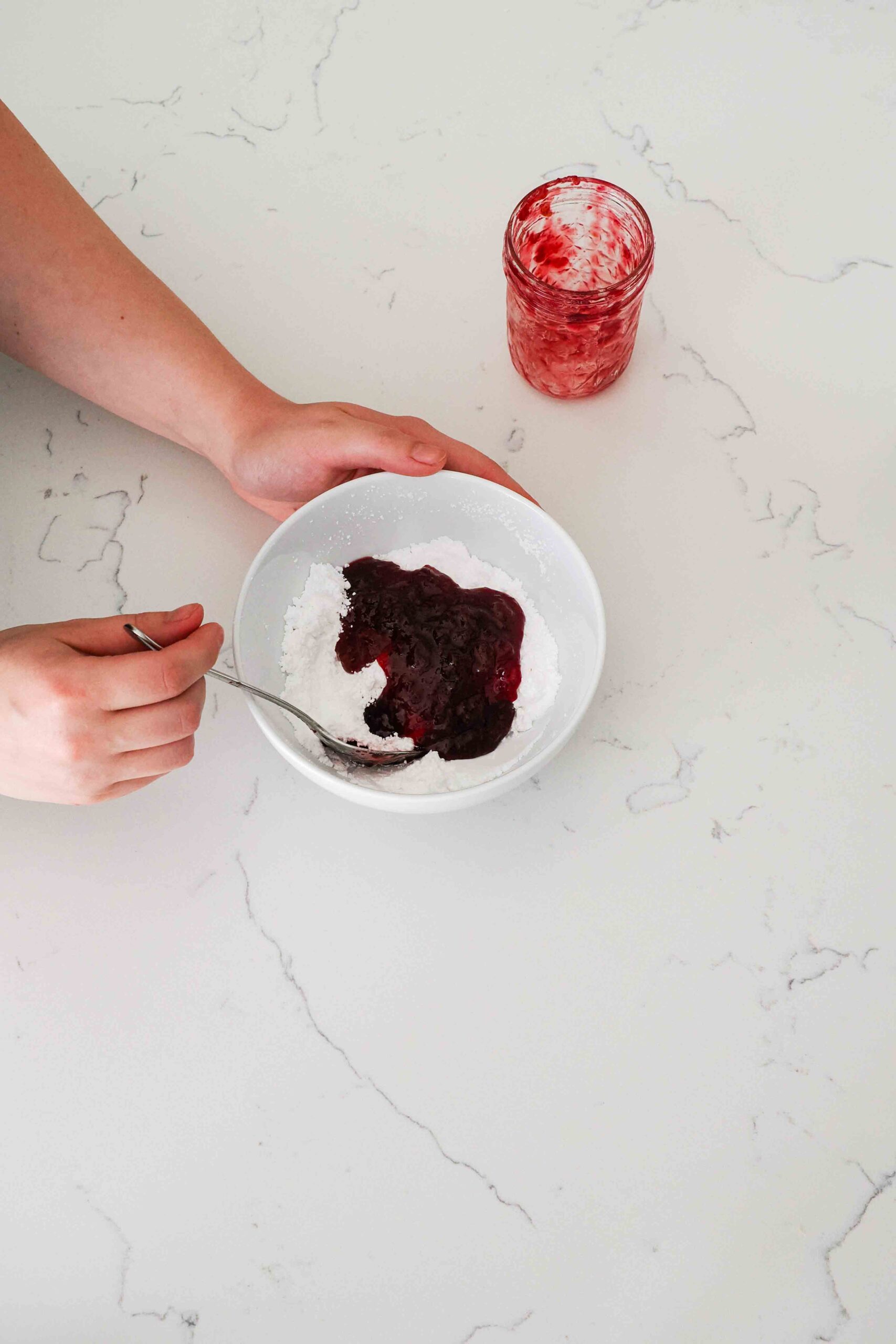 Two hands begin stirring together cherry jam and powdered sugar in a white bowl.