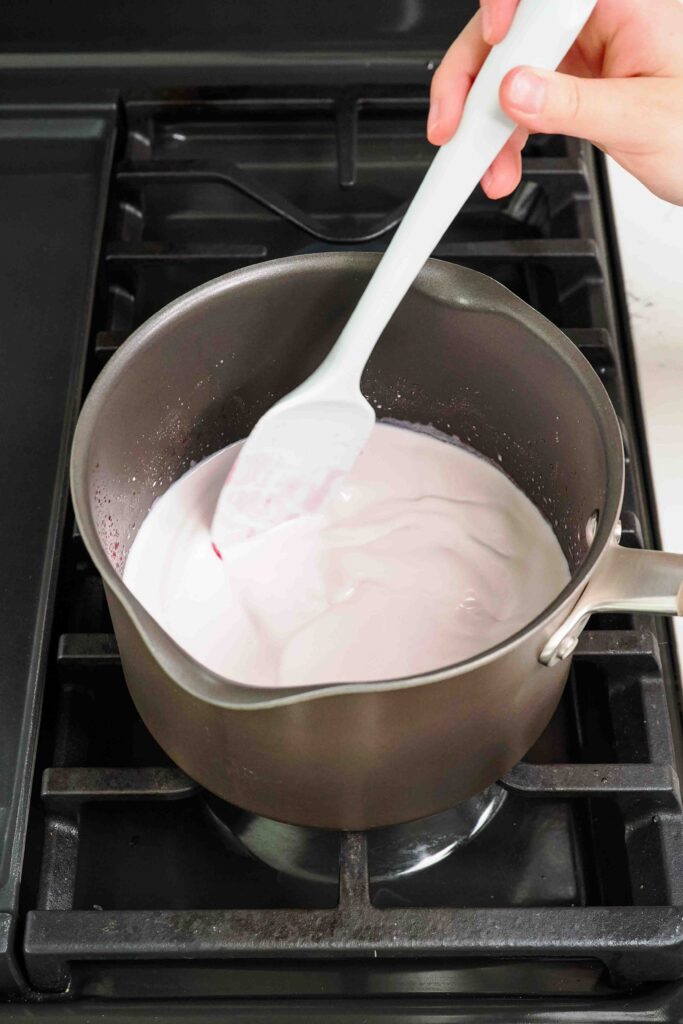 A spatula stirs milk, cream, and cherry syrup together in a pot.