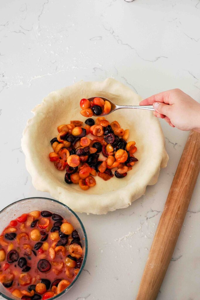 A hand spoons cherries into pie crust in a pie pan.