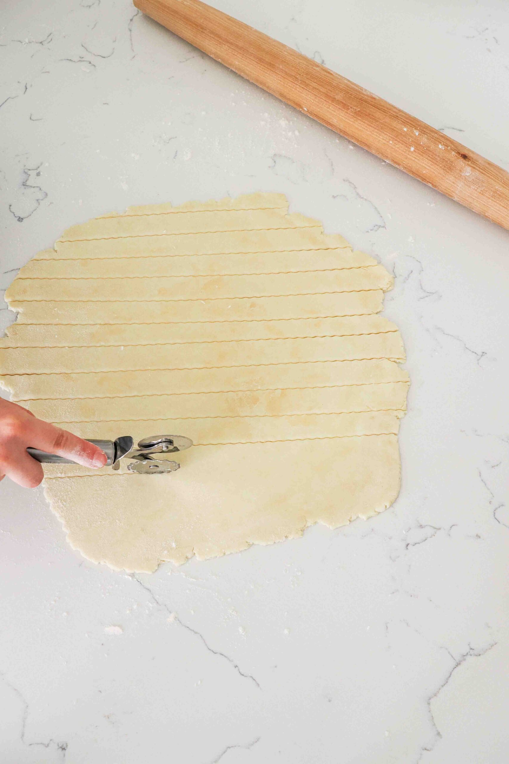 A hand uses a pastry roller to cut strips in pie dough.