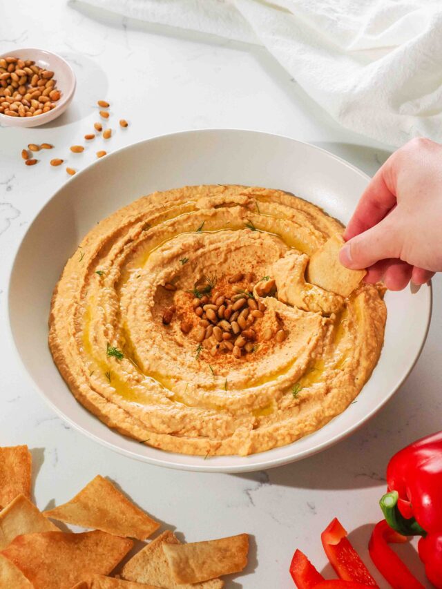 A hand dips a pita chip into a low bowl of orange hummus.