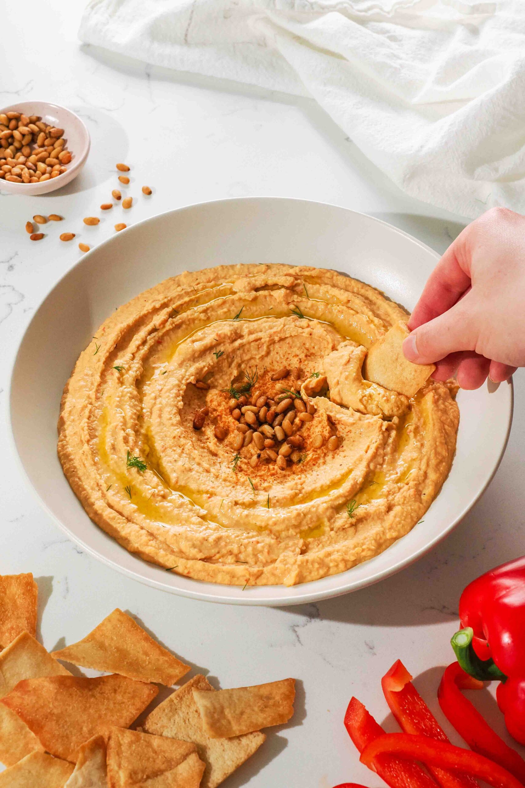 A hand dips a pita chip into a low bowl of orange hummus.